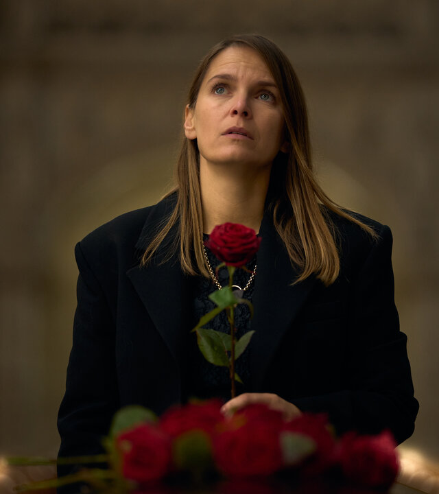 "Elixir – The Pain Killers - Das Gehirn": Isabelle (Hanna Verboom) steht in einer Kirche. Sie ist schwarz gekleidet, schaut nach oben und hält eine rote Rose in der Hand. Vor ihr liegt der Deckel eines Sargs, auf dem weitere Rosen liegen.