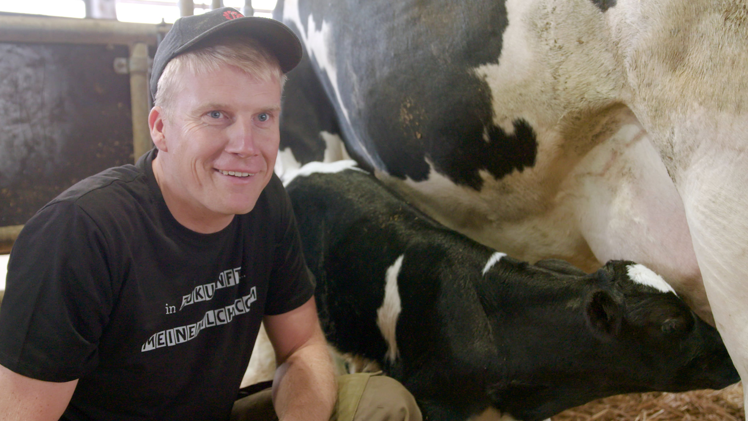 "plan b - Die gute Milch": Milchbauer Frank Lenz kniet im Stall neben einem Kälbchen, das bei seiner Mutter trinkt.
