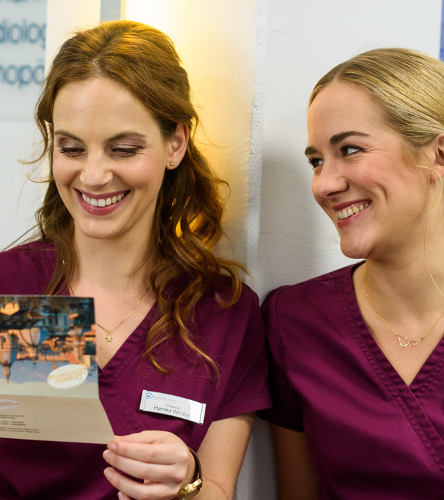 "Bettys Diagnose - Konsequenzen": Hanna (Marie Zielcke) hält eine Einladungskarte nach Rom in ihren Händen, die Betty (Annina Hellenthal) von ihrem Verehrer erhalten hat.