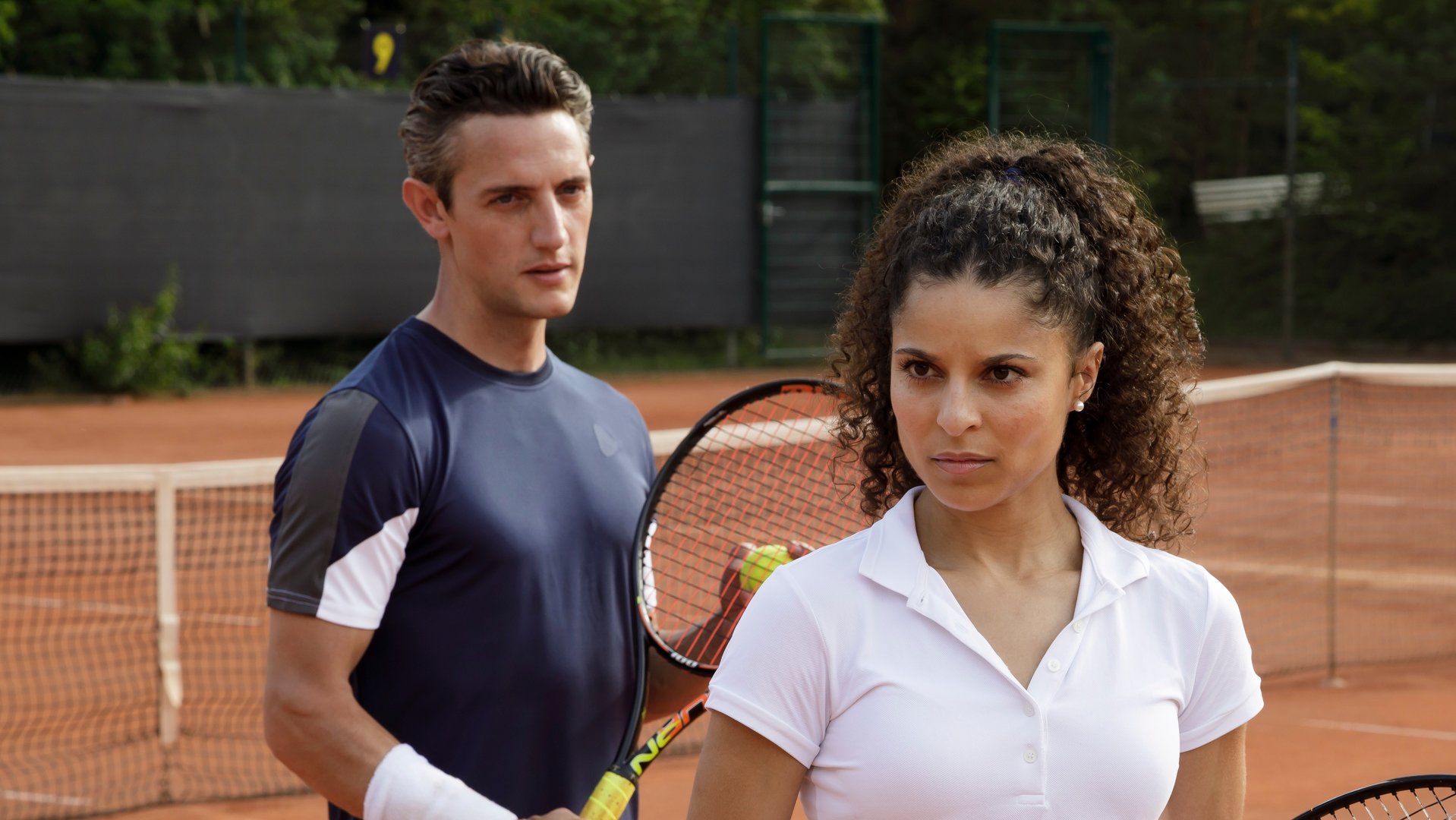 "SOKO Potsdam - Matchball": Alex Grigorov (Adam Bay) steht auf dem Tennisplatz hinter Karolin Rothe (Patricia Meeden) und schaut zu ihr. Beide halten Tennisschläger in ihren Händen. Sie blickt konzentriert nach vorne.