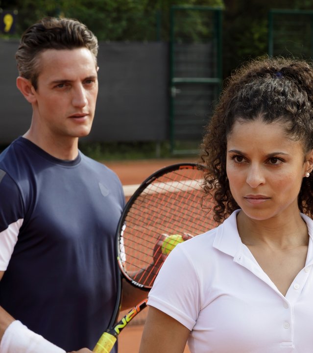 "SOKO Potsdam - Matchball": Alex Grigorov (Adam Bay) steht auf dem Tennisplatz hinter Karolin Rothe (Patricia Meeden) und schaut zu ihr. Beide halten Tennisschläger in ihren Händen. Sie blickt konzentriert nach vorne.