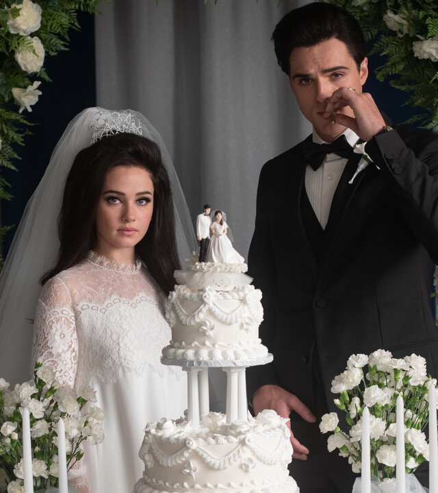 "Priscilla": Halbnahe: Priscilla (Cailee Spaeny) und Elvis (Jacob Elordi) posieren hinter einer großen Torte mit Figuren von ihnen selbst als Deko und vor einem Bogen aus weißen Rosen mit ernsten Blicken für ein Hochzeitsfoto. Elvis, der einen Smoking trägt, zieht dabei an einer Zigarette.