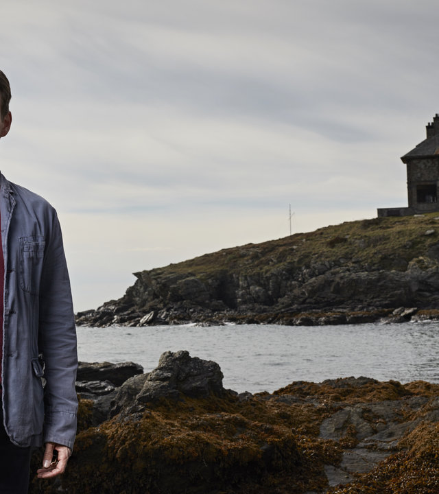 "Safe House - Entführung": Tom Brook (Stephen Moyer) steht vor einer Küste und schaut ernst in die Kamera. Im Hintergrund ist ein großes Haus zu sehen.