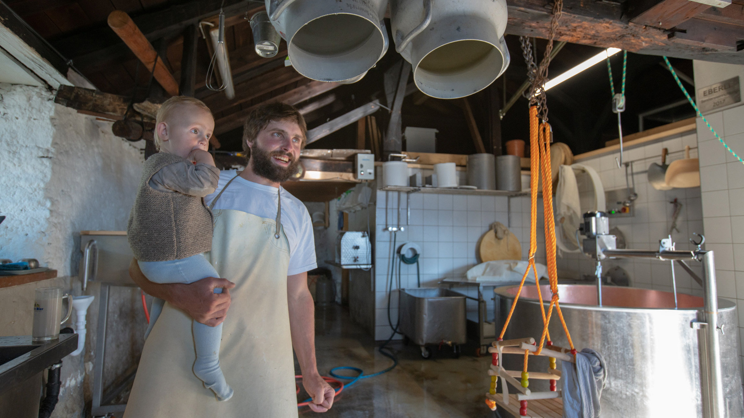 "Im Berg dahuim": Ein Mann mit Kleinkind auf dem Arm steht in einer Käserei.
