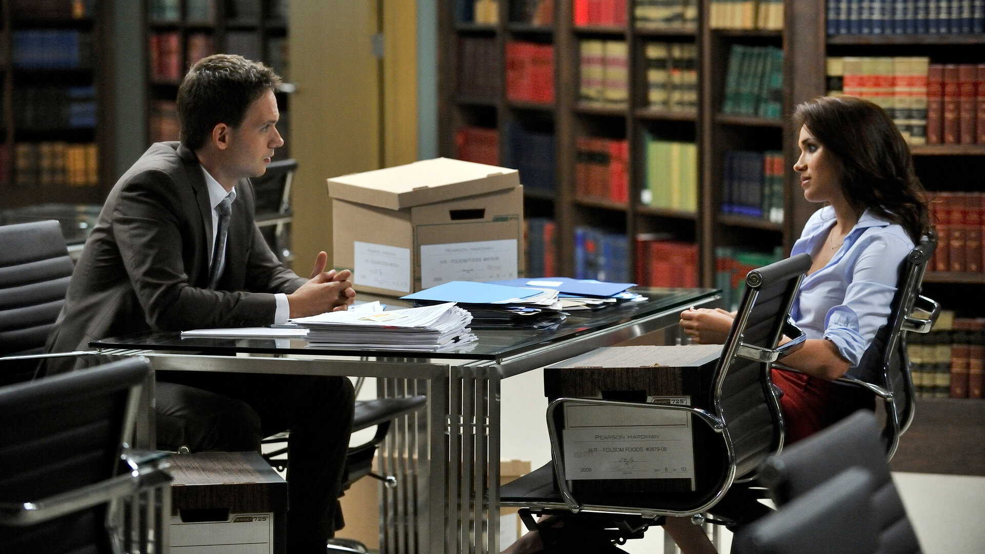 "Suits - Zane vs. Zane": Mike (Patrick J. Adams) und Rachel (Meghan Markle) sitzen sich in einem Kanzleiraum gegenüber. Auf dem Tisch vor ihnen liegen zahlreiche Akten. Beide schauen sich aufmerksam an.