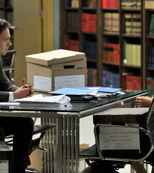 "Suits - Zane vs. Zane": Mike (Patrick J. Adams) und Rachel (Meghan Markle) sitzen sich in einem Kanzleiraum gegenüber. Auf dem Tisch vor ihnen liegen zahlreiche Akten. Beide schauen sich aufmerksam an.