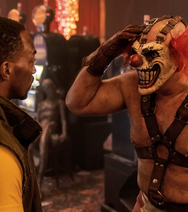 "Twisted Metal - 3RNCRCS": John (Anthony Mackie) steht Sweet Tooth (Joe Seanoa) gegenüber, der sich mit einem braunen Lederhandschuh an den Kopf fasst. Im Hintergrund sind Spielautomaten zu erkennen.