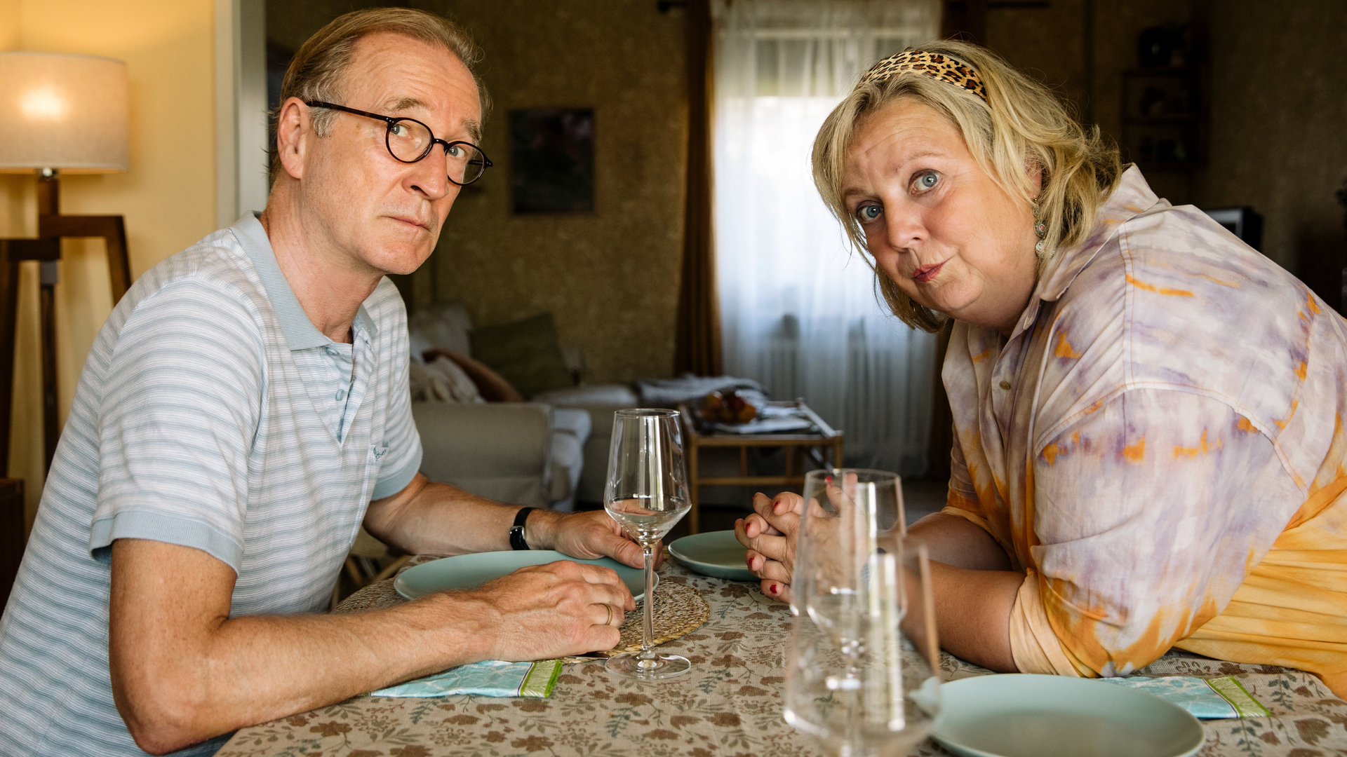 "I don't work here - Homeoffice": Heidi (Gabriela Maria Schmeide) und Walter (Peter Lohmeyer) sitzen sich am gedeckten Tisch gegenüber. Heidi hat die Unterarme auf dem Tisch abgestützt, lehnt sich nach vorne und schaut zu ihrer linken Seite. Auch Walter blickt durch seine runde Brille etwas ängstlich zur Seite und ist dabei leicht nach vorne geneigt. Seinen hellblauen Teller hält er an den Seiten fest.