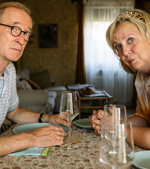 "I don't work here - Homeoffice": Heidi (Gabriela Maria Schmeide) und Walter (Peter Lohmeyer) sitzen sich am gedeckten Tisch gegenüber. Heidi hat die Unterarme auf dem Tisch abgestützt, lehnt sich nach vorne und schaut zu ihrer linken Seite. Auch Walter blickt durch seine runde Brille etwas ängstlich zur Seite und ist dabei leicht nach vorne geneigt. Seinen hellblauen Teller hält er an den Seiten fest.