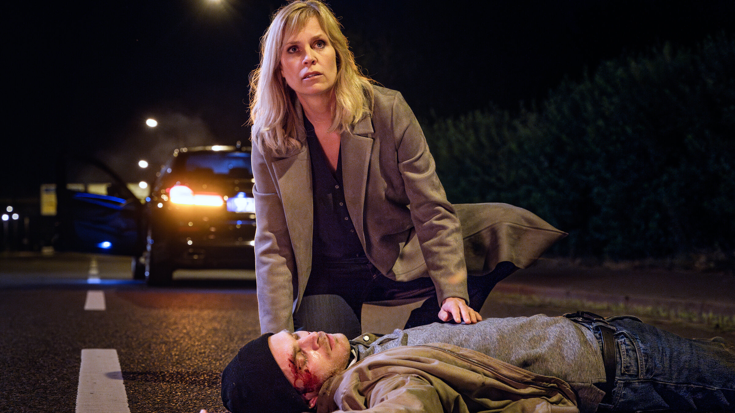 "SOKO Leipzig - Plan C": Es ist Nacht. Ina (Melanie Marschke) kniet auf einer Straße über dem bewusstlosen Jonas Stein (Leonard Proxauf). Der junge Mann hat eine Platzwunde am Kopf. Hinter Ina steht ein Auto mit geöffneter Fahrertüre.