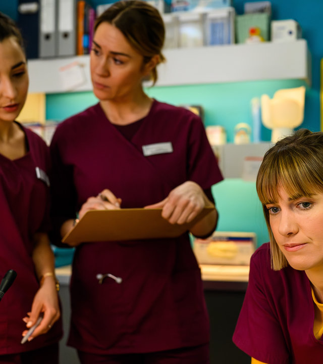 "Bettys Diagnose - Wunder geschehen": Ava (Rona Özkan) und Betty (Henrike Hahn) stehen nebeneinander und unterhalten sich. Neben ihnen sitzt Rike (Isabell Horn), die gedankenverloren geradeaus blickt. Vor ihnen steht ein Blumenstrauß in einer hellen Vase.