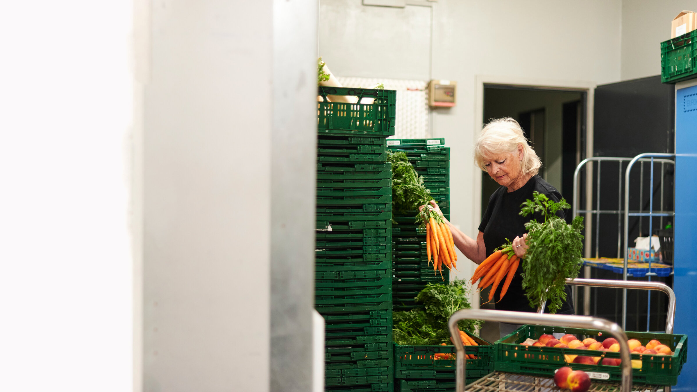 "37°: Rente? Reicht nicht!": Angelika steht in einem Lagerraum vor grünen, gestapelten Gemüsekisten. Sie hält zwei Bund Möhren in der Hand. Neben ihr steht ein Wagen mit einer Kiste mit Pfirsichen.