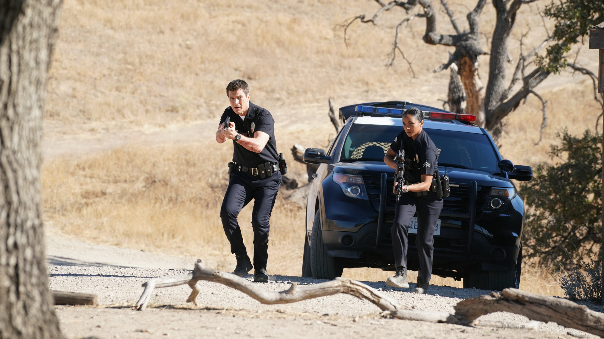"The Rookie - Tag des Todes": Schwer bewaffnet stürmen John Nolan (Nathan Fillion) und Nyla Harper (Mekia Cox) aus dem Polizeiauto auf das Grundstück in der Wüste.