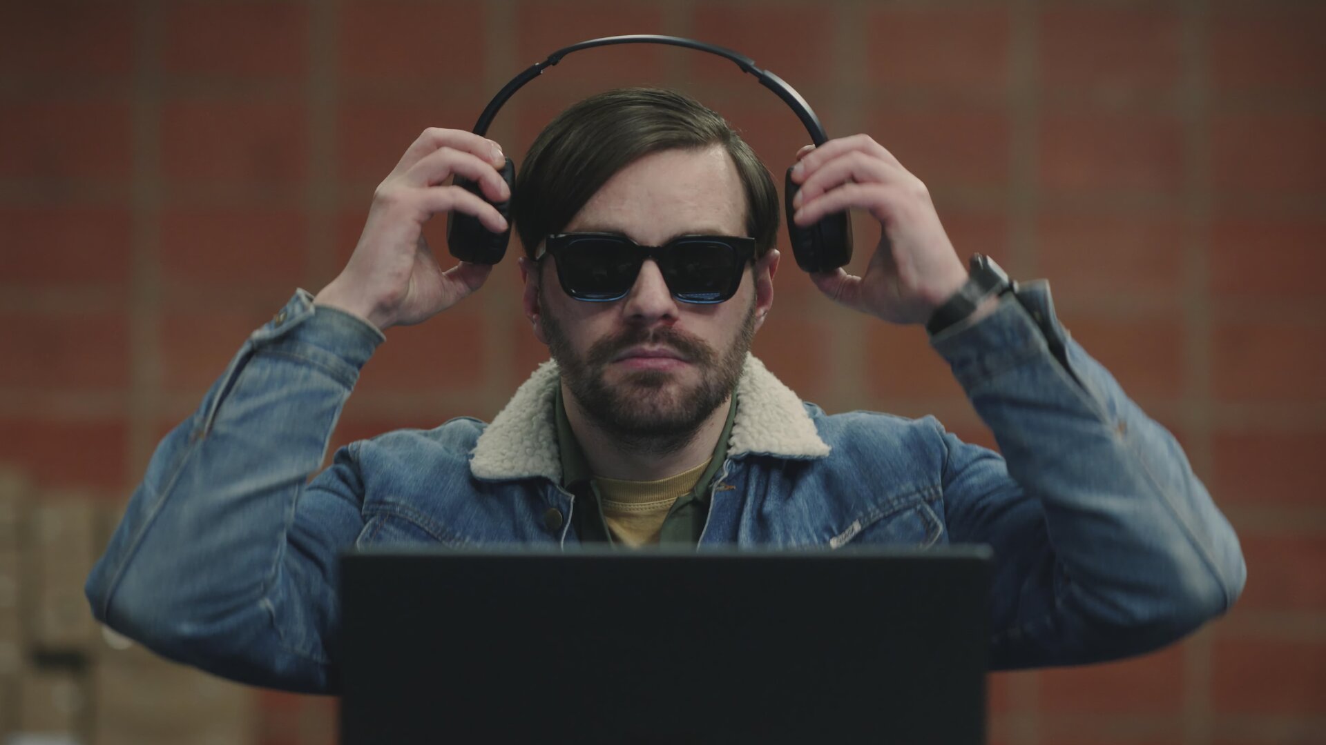 "Blind Sherlock - Ich höre was, was du nicht hörst": Roman Mertens (Bart Kelchtermans) sitzt in einem Raum vor einem aufgeklappten Laptop. Er trägt eine dunkle Brille und setzt sich gerade Kopfhörer auf.