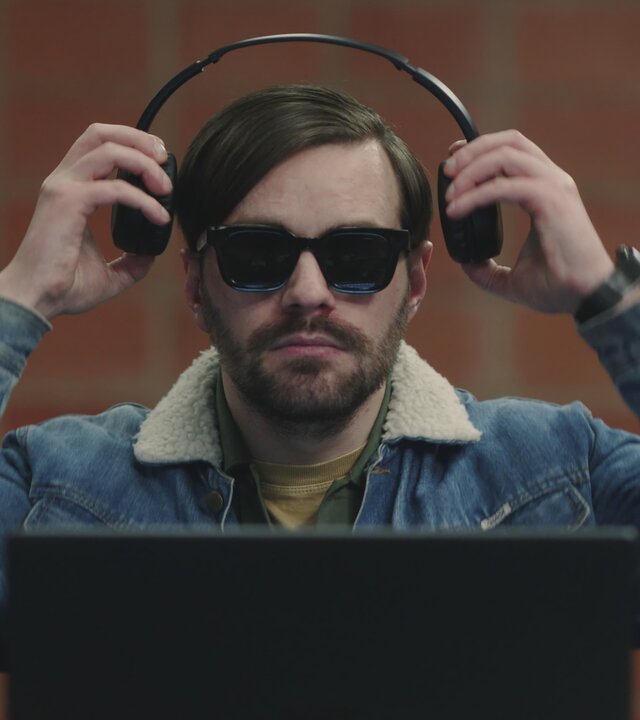 "Blind Sherlock - Ich höre was, was du nicht hörst": Roman Mertens (Bart Kelchtermans) sitzt in einem Raum vor einem aufgeklappten Laptop. Er trägt eine dunkle Brille und setzt sich gerade Kopfhörer auf.