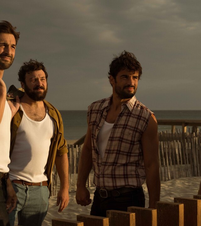 "Escort Boys - Imkerei mit Zusatzleistungen": Ben (Guillaume Labbé), Ludo (Thibaut Evrard), Mathias (Simon Ehrlacher) und Zak (Corentin Fila) stehen vor einem braunen Holzzaun am Meer. Ben und Zak haben große Taschen bei sich: Ben trägt seine Tasche über der linken Schulter, Zak hat seine auf dem Zaun abgestellt und stützt sich mit verschränkten Armen darauf. Alle vier tragen leichte Kleidung und blicken konzentriert. Sonnenlicht blendet ihre Gesichter leicht.