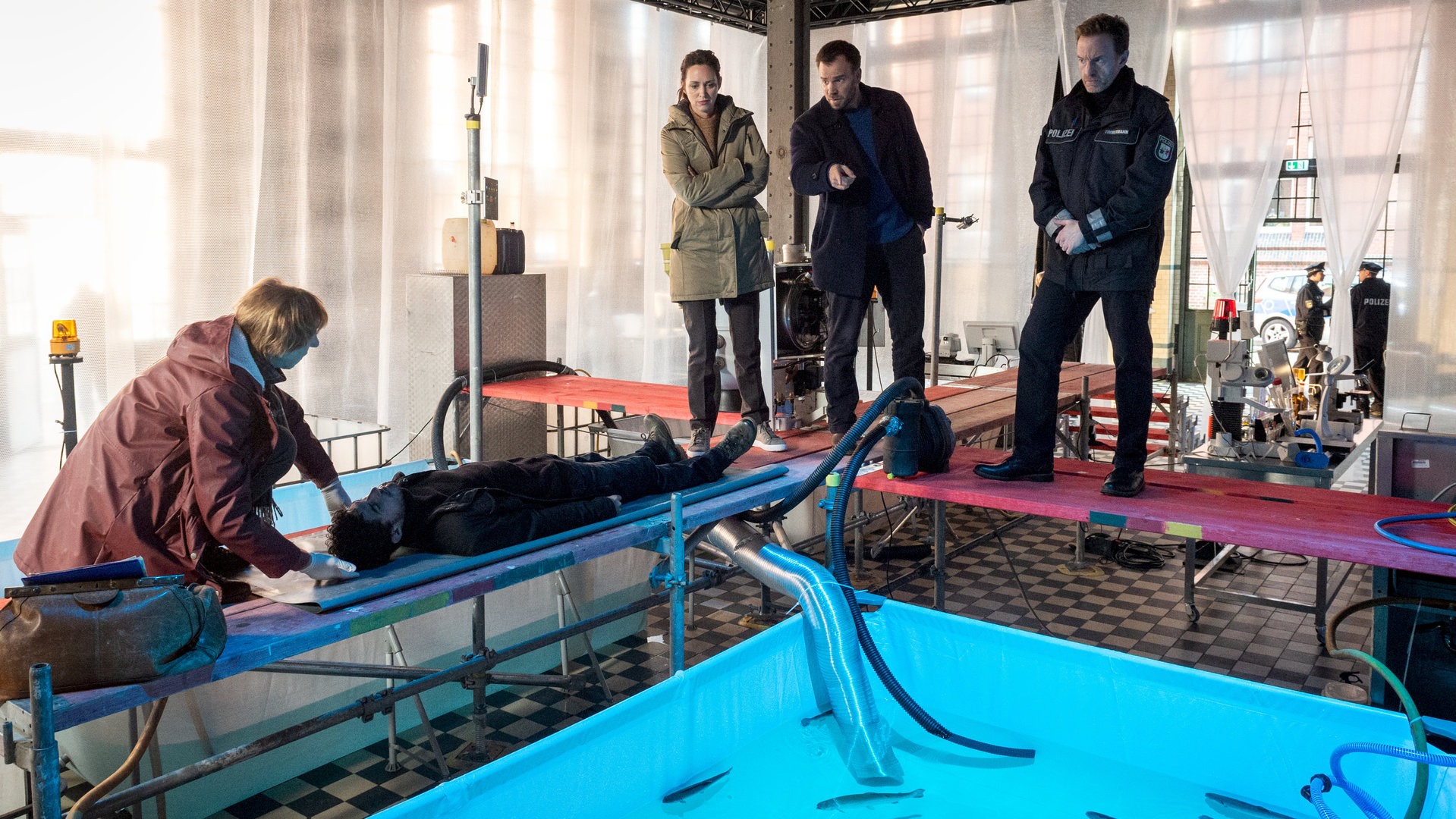 "SOKO Wismar – Die Rache der Ostseeschnäpel": In einer Fischzuchtanlage schwimmen in einem Wasserbecken Fische. Ringsum befinden sich Stege. Links auf einem Steg liegt der Tote (Alexander Berenheuser). An seinem Kopfende hockt Helen (Katharina Blaschke). Auf einem anderen Steg stehen Timmermann (Mathias Junge) und die Kommissare Joost (Nike Fuhrmann) und Pöhlmann (Dominic Boeer). Alle blicken zum Toten.