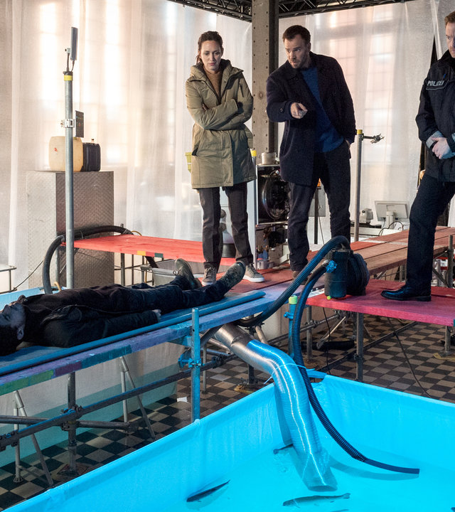 "SOKO Wismar – Die Rache der Ostseeschnäpel": In einer Fischzuchtanlage schwimmen in einem Wasserbecken Fische. Ringsum befinden sich Stege. Links auf einem Steg liegt der Tote (Alexander Berenheuser). An seinem Kopfende hockt Helen (Katharina Blaschke). Auf einem anderen Steg stehen Timmermann (Mathias Junge) und die Kommissare Joost (Nike Fuhrmann) und Pöhlmann (Dominic Boeer). Alle blicken zum Toten.