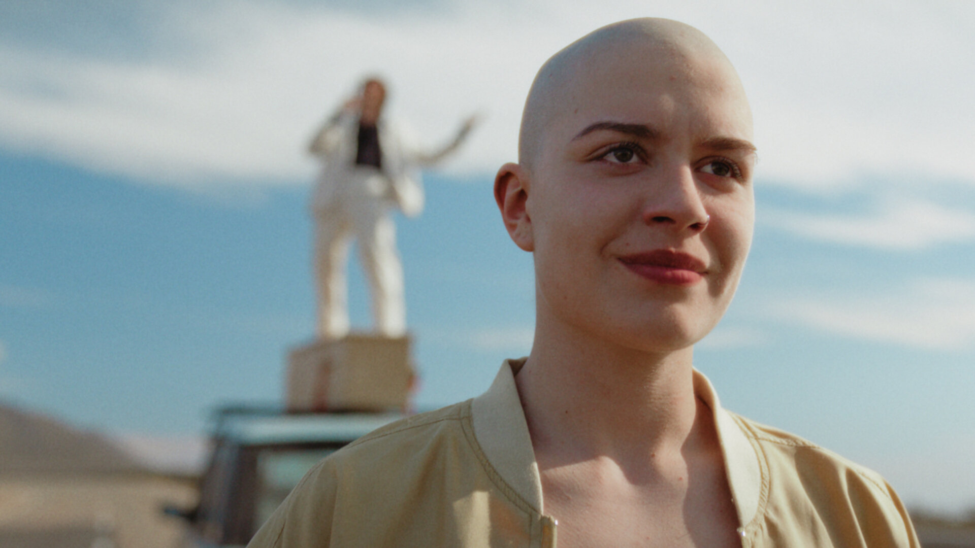 "How to Kill Your Sister - Folge 5": Die kahlköpfige Anna (Emma Rotsaert) steht auf einer Straße. Sie lächelt. Im Hintergrund ist unscharf ein altes Auto mit einem Holzsarg auf dem Dach zu sehen. Darauf steht ein weiß gekleideter Mann und singt.