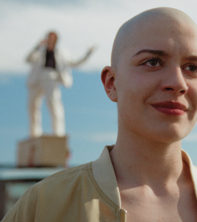 "How to Kill Your Sister - Folge 5": Die kahlköpfige Anna (Emma Rotsaert) steht auf einer Straße. Sie lächelt. Im Hintergrund ist unscharf ein altes Auto mit einem Holzsarg auf dem Dach zu sehen. Darauf steht ein weiß gekleideter Mann und singt.