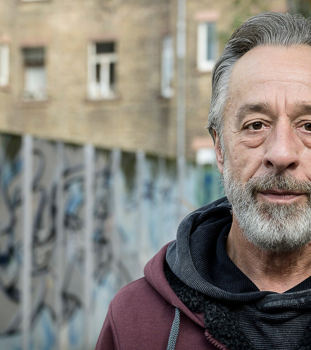 "Zwei Quadratkilometer Stress - Hilfe für einen Stadtteil": Stefan Memel, Sozialarbeiter und Gründer von Aufwind-Mannheim e.V. , steht vor einer mit Graffiti besprühten Mauer in der Mannheimer Neckarstadt und schaut in die Kamera.