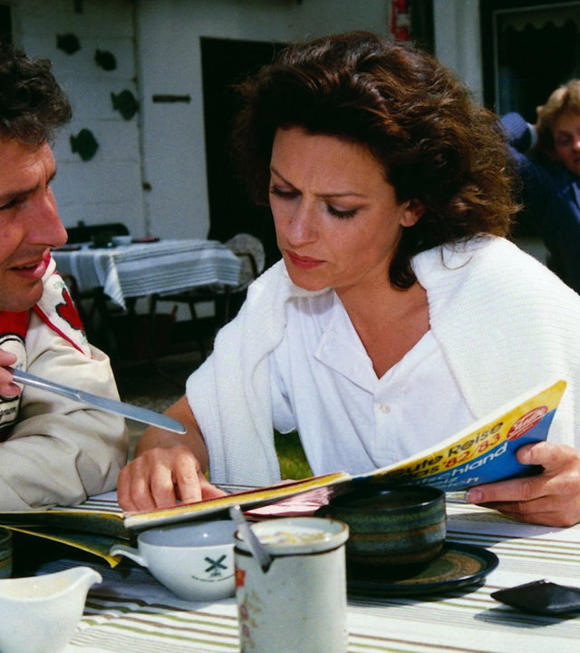 "Der Landarzt - Das Attentat": Corinna Martens (Beatrice Kessler) und ihr Rallye-Kopilot (Volker Brandt) sitzen bei einer Tasse Kaffee am Tisch. Sie studiert eine Straßenkarte, er schwingt sein Brotmesser.