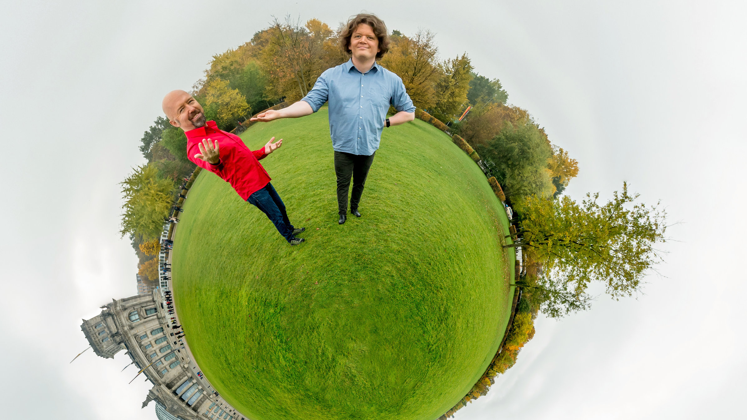 "Der satirische Jahresrückblick 2019": Die Satire-Autoren Andreas Wiemers und Werner Doyé stehen auf einer mit Gras bedeckten Kugel und schauen in die Kamera. Neben ihnen ist das Reichstagsgebäude zu sehen, hinter ihnen Bäume. Die beiden Männer sind im Verhältnis zu ihrer Umgebung riesengroß.