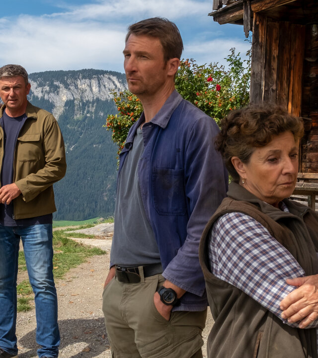 "Der Bergdoktor: Spiel mit dem Feuer (2)": Auf dem Gruberhof steht Martin (Hans Sigl) abseits mit gepackter Tasche über der Schulter tragend und schaut zu Lisbeth (Monika Baumgartner) und Hans (Heiko Ruprecht). Beide stehen ihm abgewandt und blicken ernst zur Seite.