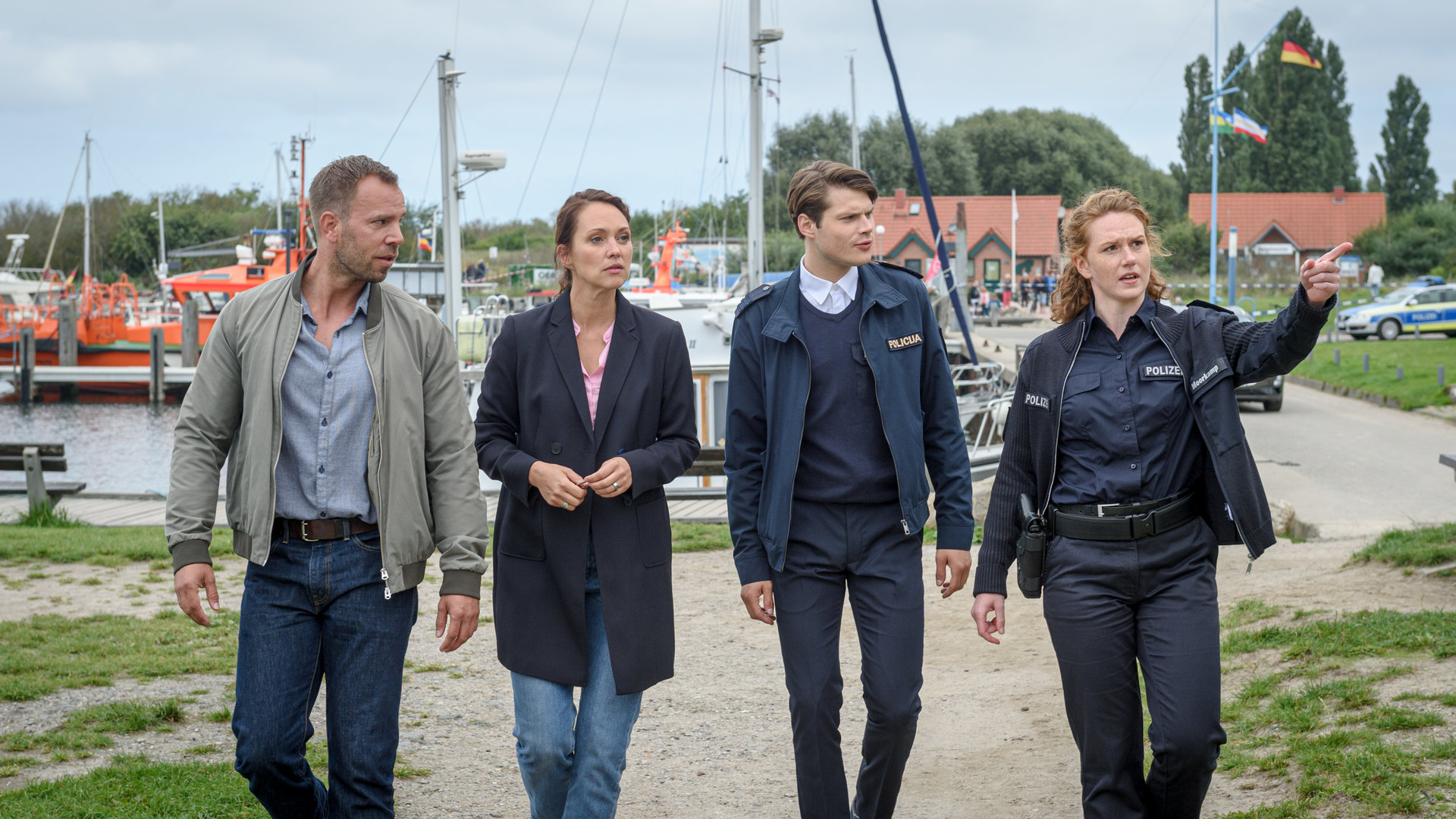 "SOKO Wismar – Ein bürokratischer Tod": An einem Bootshafen in der Nähe von Wismar gehen die Kommissare Karoline Joost (Nike Fuhrmann) und Lars Pöhlmann (Dominic Boeer) sowie die Polizistin Paula Moorkamp (Stella Hinrichs) und ihr neuer Kollege Eddi Jansons (Gustavs Gailus) in Richtung des Tatorts. Paula zeigt auf etwas außerhalb des Bildes, hinter ihnen sind Anleger mit mehreren Booten zu sehen.