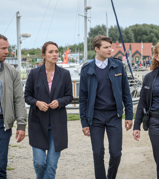 "SOKO Wismar – Ein bürokratischer Tod": An einem Bootshafen in der Nähe von Wismar gehen die Kommissare Karoline Joost (Nike Fuhrmann) und Lars Pöhlmann (Dominic Boeer) sowie die Polizistin Paula Moorkamp (Stella Hinrichs) und ihr neuer Kollege Eddi Jansons (Gustavs Gailus) in Richtung des Tatorts. Paula zeigt auf etwas außerhalb des Bildes, hinter ihnen sind Anleger mit mehreren Booten zu sehen.