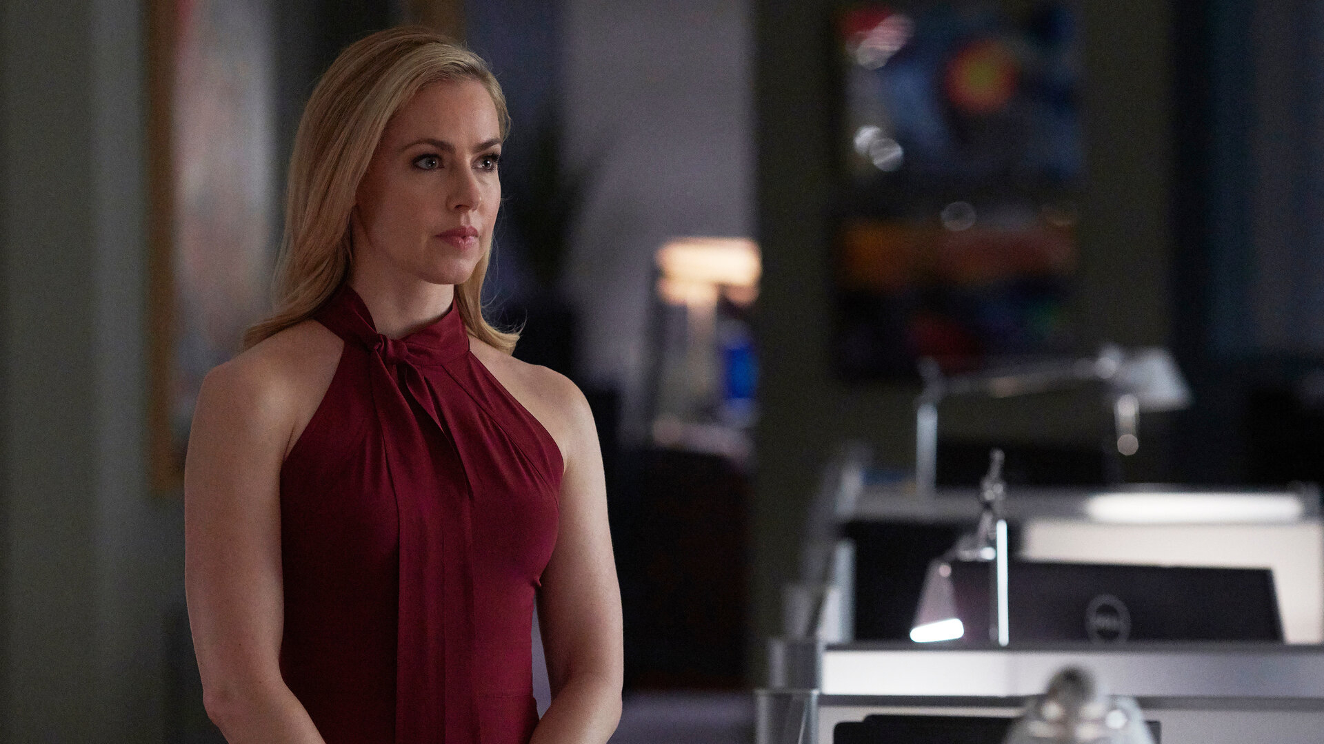 "Suits - Das große Ganze": Katrina (Amanda Schull) trägt ein dunkelrotes Kleid und schaut nachdenklich. Sie steht im Großraumbüro der Kanzlei, neben ihr sind Monitore und Schreibtischlampen zu sehen.