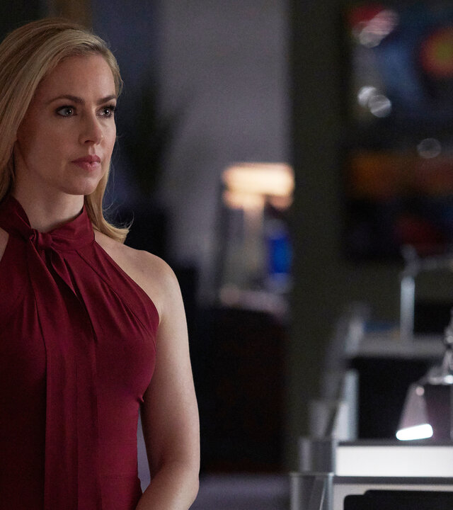 "Suits - Das große Ganze": Katrina (Amanda Schull) trägt ein dunkelrotes Kleid und schaut nachdenklich. Sie steht im Großraumbüro der Kanzlei, neben ihr sind Monitore und Schreibtischlampen zu sehen.