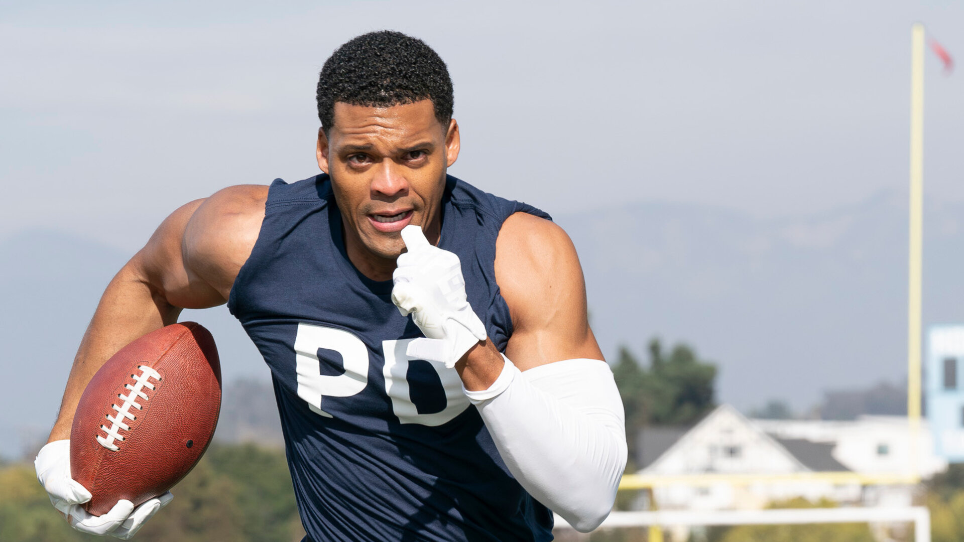 "The Rookie - Mad about Murder": Miles Penn (Deric Augustine) sprintet über ein Footballfeld und hält den Ball fest in der Hand. Er trägt dunkle Sportkleidung: ein dunkelblaues Tanktop, eine schwarze Hose mit weißen und blauen Details, außerdem weiße Handschuhe, einen weißen Arm Sleeve und eine schwarze Knieschiene. Sein Blick ist konzentriert nach vorn gerichtet. Hinter ihm läuft ein weiterer Mitspieler (Jan M. Janssen), der versucht, aufzuschließen. Im Hintergrund befinden sich weitere Spieler (Komparsen) in roten Trikots.