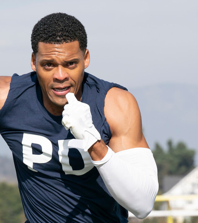 "The Rookie - Mad about Murder": Miles Penn (Deric Augustine) sprintet über ein Footballfeld und hält den Ball fest in der Hand. Er trägt dunkle Sportkleidung: ein dunkelblaues Tanktop, eine schwarze Hose mit weißen und blauen Details, außerdem weiße Handschuhe, einen weißen Arm Sleeve und eine schwarze Knieschiene. Sein Blick ist konzentriert nach vorn gerichtet. Hinter ihm läuft ein weiterer Mitspieler (Jan M. Janssen), der versucht, aufzuschließen. Im Hintergrund befinden sich weitere Spieler (Komparsen) in roten Trikots.