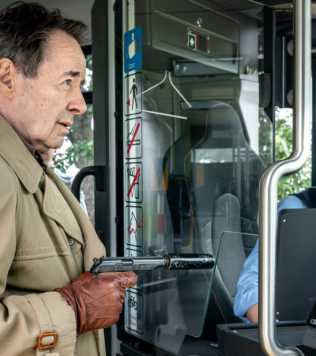 "SOKO Leipzig - Blinde Flecken": Ein Mann (Uwe Kockisch) betritt einen Bus, er trägt Lederhandschuhe und einen Trenchcoat. Währenddessen hält er der Busfahrerin (Maria Brendel) eine Waffe vor.