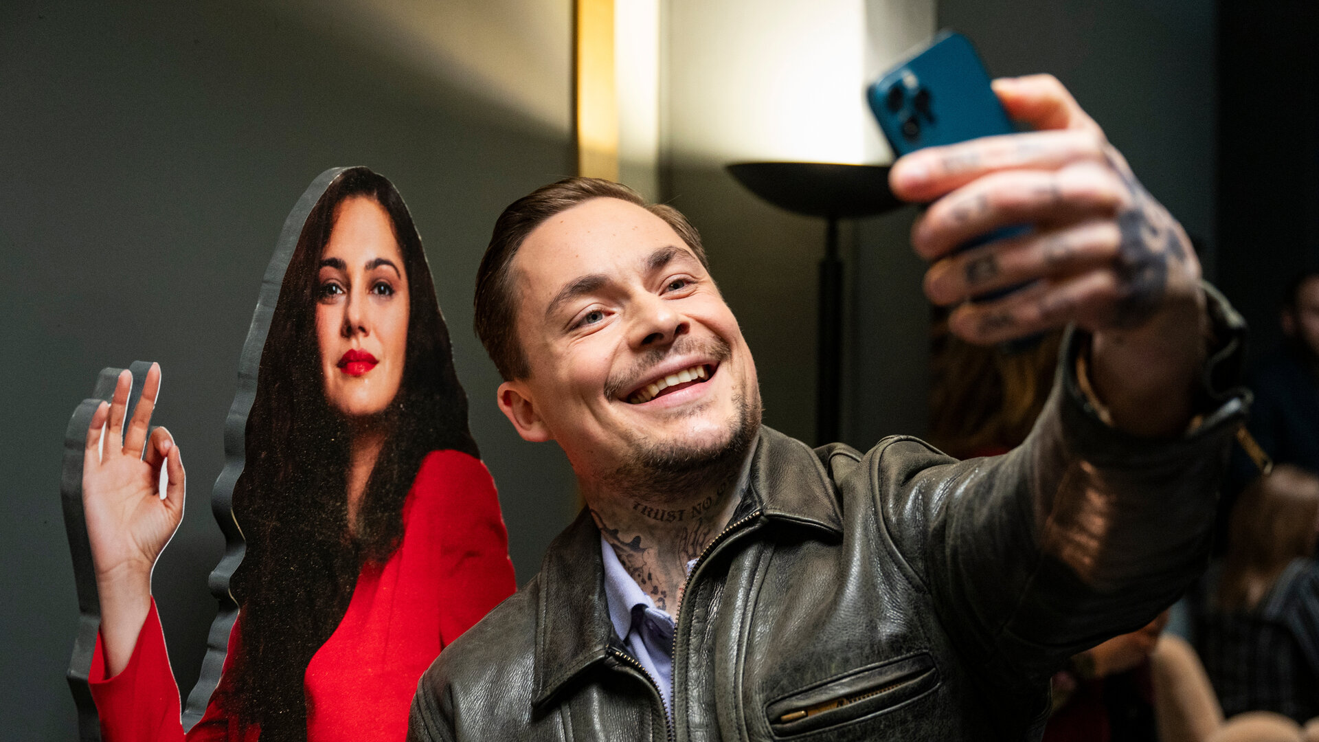 "Take the Money and Run - One Family": Nikolaj Ignatov (Ferdinand Lehmann) macht in brauner Lederjacke ein Selfie von sich und einem Papp-Aufsteller von Ruja Ignatova (Nilam Farooq).