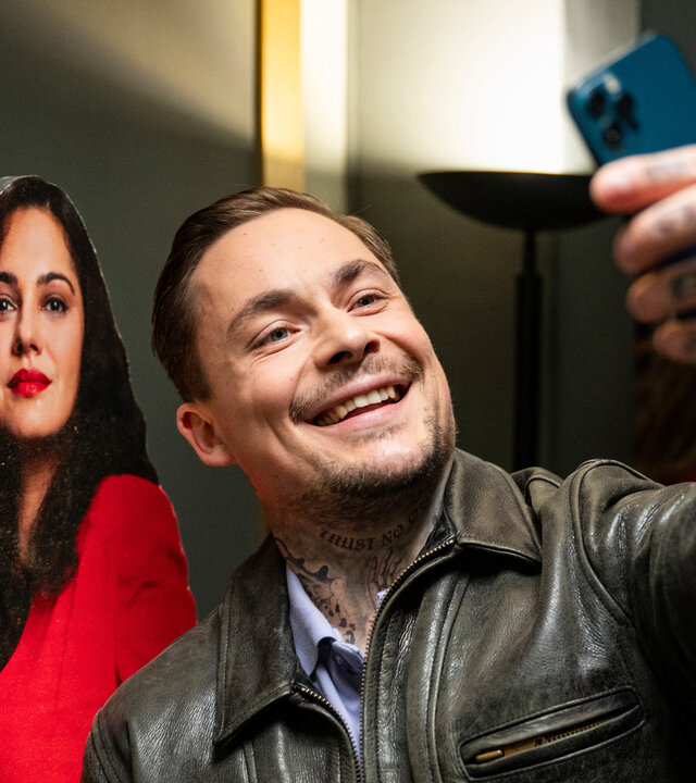 "Take the Money and Run - One Family": Nikolaj Ignatov (Ferdinand Lehmann) macht in brauner Lederjacke ein Selfie von sich und einem Papp-Aufsteller von Ruja Ignatova (Nilam Farooq).
