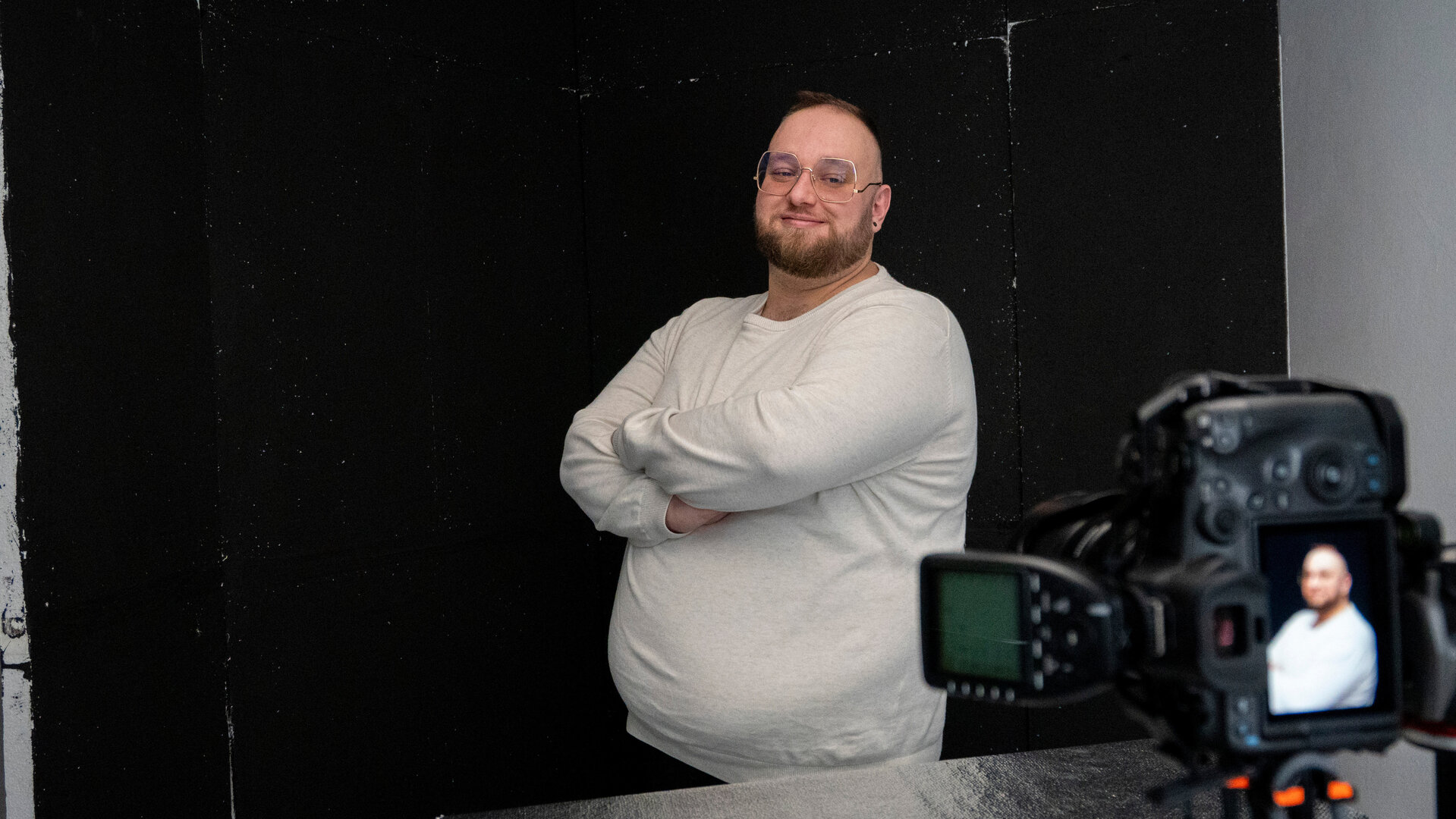 "einfach Mensch - Stefan Rosenberger: Body Positivity (2)": An der Seite halten Hände eine Kamera. Dahinter steht Stefan Rosenberger vor einem dunklem Hintergrund zur Seite gedreht und lächelt in Richtung Kamera. Er hat die Arme verschränkt.