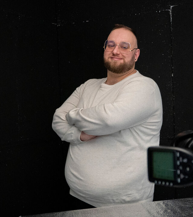 "einfach Mensch - Stefan Rosenberger: Body Positivity (2)": An der Seite halten Hände eine Kamera. Dahinter steht Stefan Rosenberger vor einem dunklem Hintergrund zur Seite gedreht und lächelt in Richtung Kamera. Er hat die Arme verschränkt.