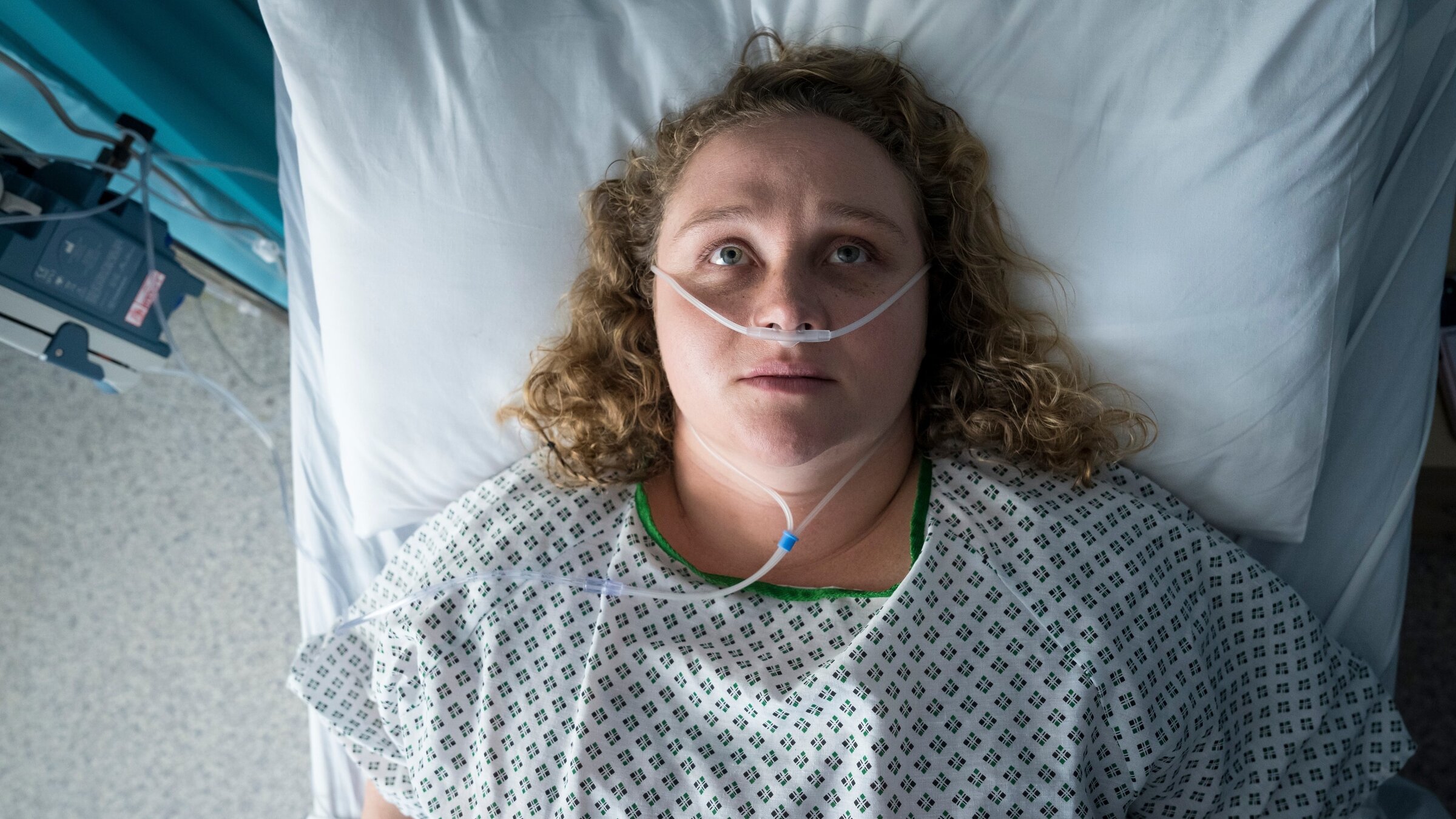 "The Tourist - Irisches Blut - In Morpheus' Fängen": Blick von oben: Helen Chambers (Danielle Macdonald) liegt in einem Krankenbett. Sie trägt ein grün-weißes Nachthemd der Klinik; in ihrer Nase hängt ein Plastikschlauch zur Versorgung mit Sauerstoff. Helen hat die Augen geöffnet und schaut nach oben an die Zimmerdecke.