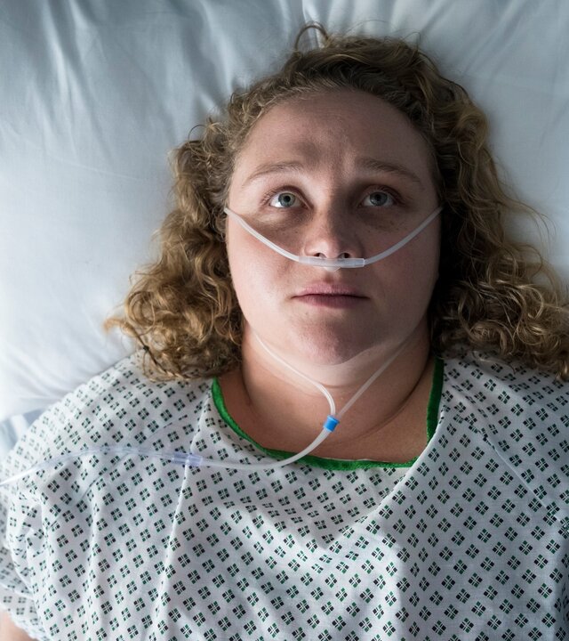 "The Tourist - Irisches Blut - In Morpheus' Fängen": Blick von oben: Helen Chambers (Danielle Macdonald) liegt in einem Krankenbett. Sie trägt ein grün-weißes Nachthemd der Klinik; in ihrer Nase hängt ein Plastikschlauch zur Versorgung mit Sauerstoff. Helen hat die Augen geöffnet und schaut nach oben an die Zimmerdecke.