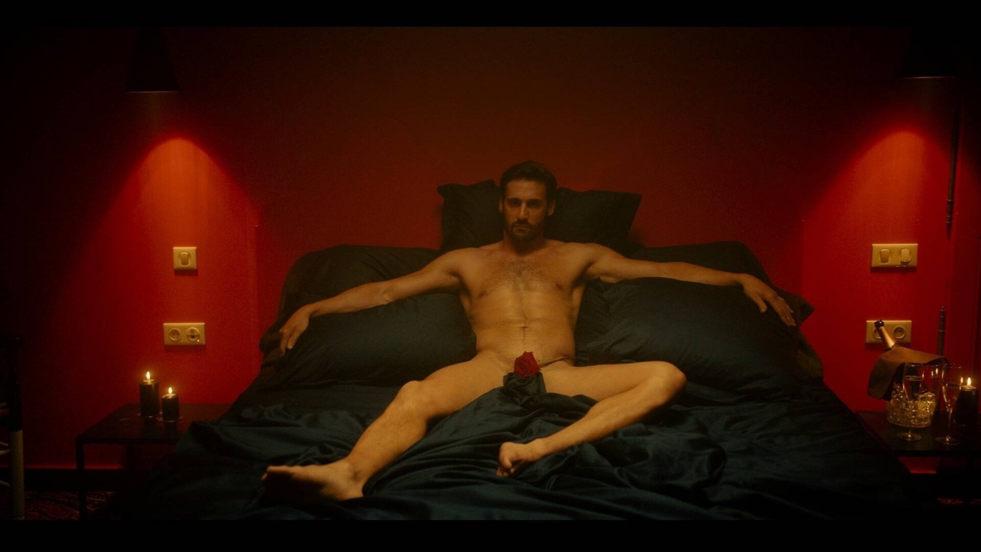 "Escort Boys - Ein letztes Mal": Ben (Guillaume Labbé) liegt nackt auf einem schwarzen Bett in einem Schlafzimmer. Eine Rose bedeckt seinen Schoß. Das Licht ist gedimmt, auf dem Nachttisch brennen Kerzen und tauchen den Raum in warmes Licht.