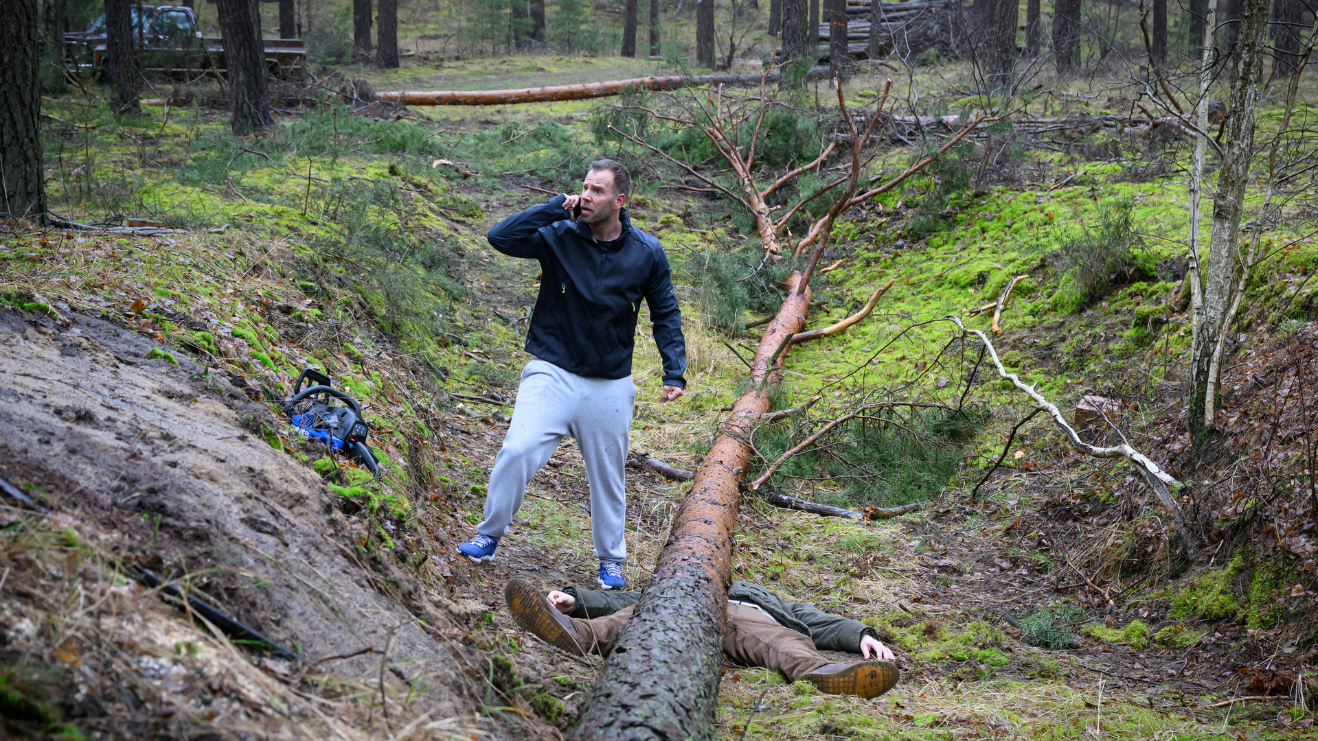 "SOKO Wismar - Holz fällt!": Lars Pöhlmann (Dominic Boeer) steht im Wald neben der Leiche von Alexander Hübner (Odysseas J. Hlepas) und telefoniert.
