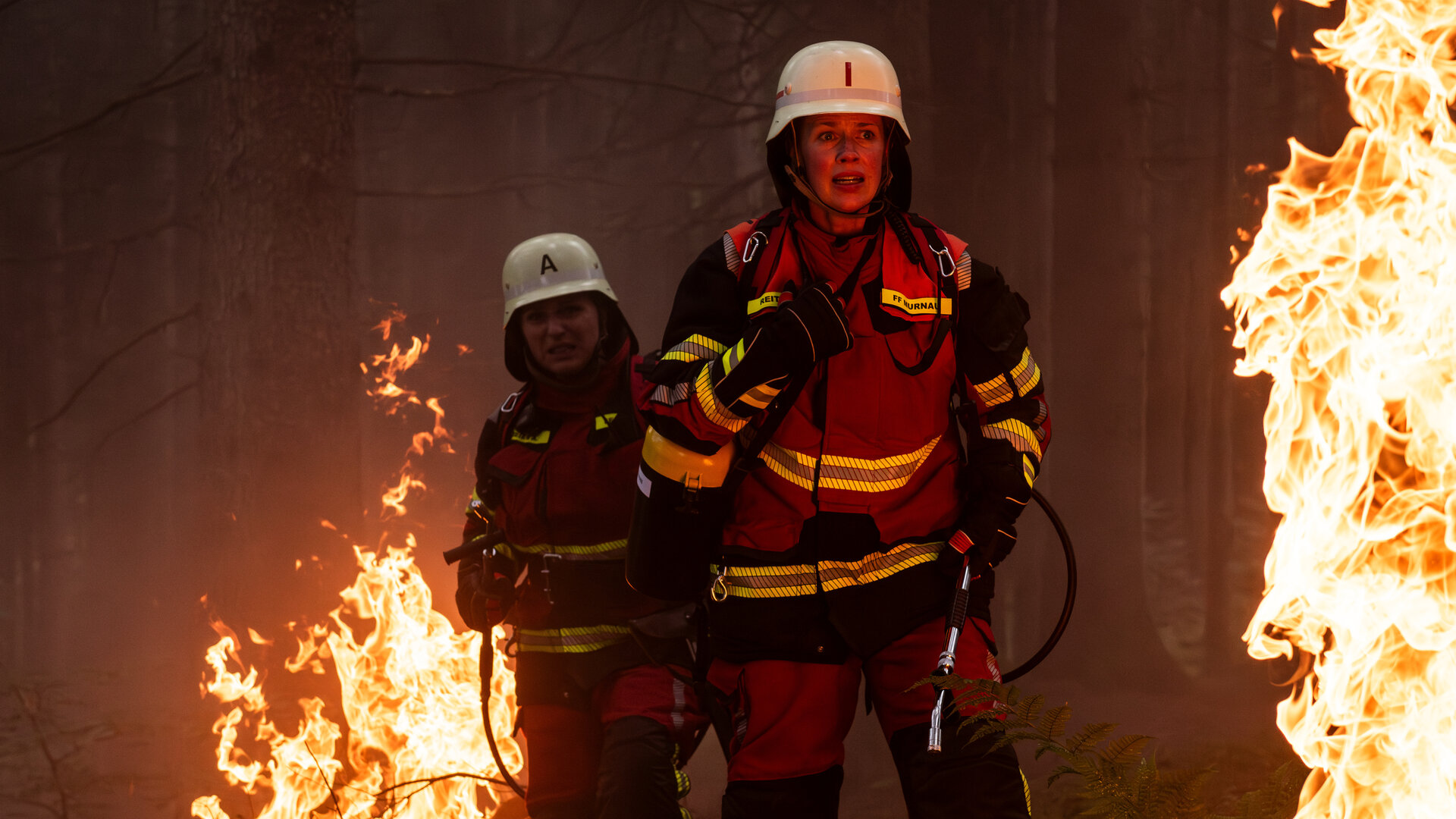 "Marie fängt Feuer - Hitzewelle": Marie (Christine Eixenberger) und Toni (Lisa Jopt) stehen in ihren Feuerwehruniformen im Wald und sind von Flammen umgeben.