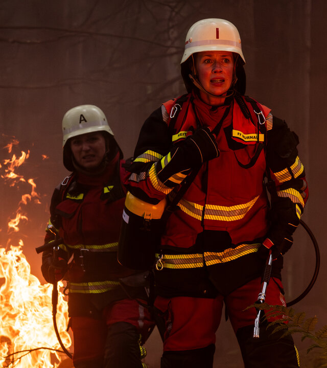 "Marie fängt Feuer - Hitzewelle": Marie (Christine Eixenberger) und Toni (Lisa Jopt) stehen in ihren Feuerwehruniformen im Wald und sind von Flammen umgeben.