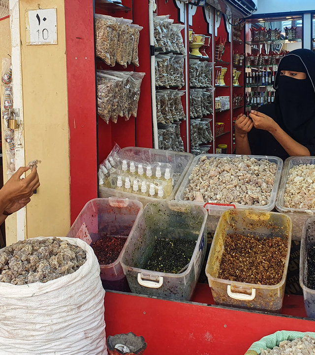 "Arabiens Traum von der Zukunft - Von Oman nach Saudi-Arabien": Golineh Atai steht lachend an einem Weihrauchstand in Oman. Sie hält etwas Weihrauch in der Hand, vor ihr steht ein großer Beutel mit Weihrauch. Die verschleierte Händlerin steht hinter mehreren Plastikbehältern mit verschiedenfarbigen Waren. Sie hält ebenfalls etwas in der Hand und hat sich Golineh Atai zugewandt.