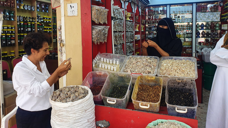 "Arabiens Traum von der Zukunft - Von Oman nach Saudi-Arabien": Golineh Atai steht lachend an einem Weihrauchstand in Oman. Sie hält etwas Weihrauch in der Hand, vor ihr steht ein großer Beutel mit Weihrauch. Die verschleierte Händlerin steht hinter mehreren Plastikbehältern mit verschiedenfarbigen Waren. Sie hält ebenfalls etwas in der Hand und hat sich Golineh Atai zugewandt.
