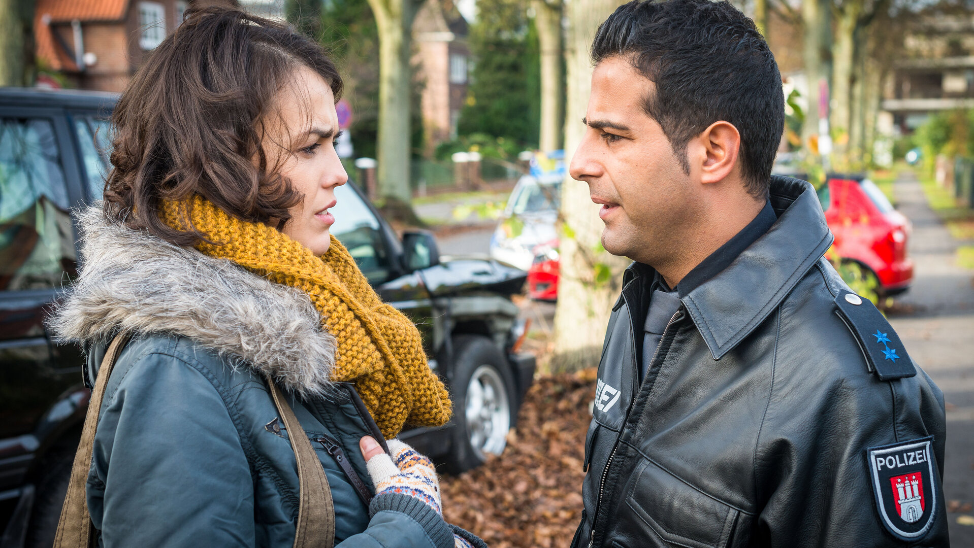 "Notruf Hafenkante - Paulines Fall": Auf einem Gehweg am Straßenrand stehen sich der Polizist Tarik Coban (Serhat Cokgezen) und Pauline Becker (Lucie Heinze) gegenüber und sehen sich an. Im Hintergrund sind parkende Autos, Bäume und Häuser zu sehen.