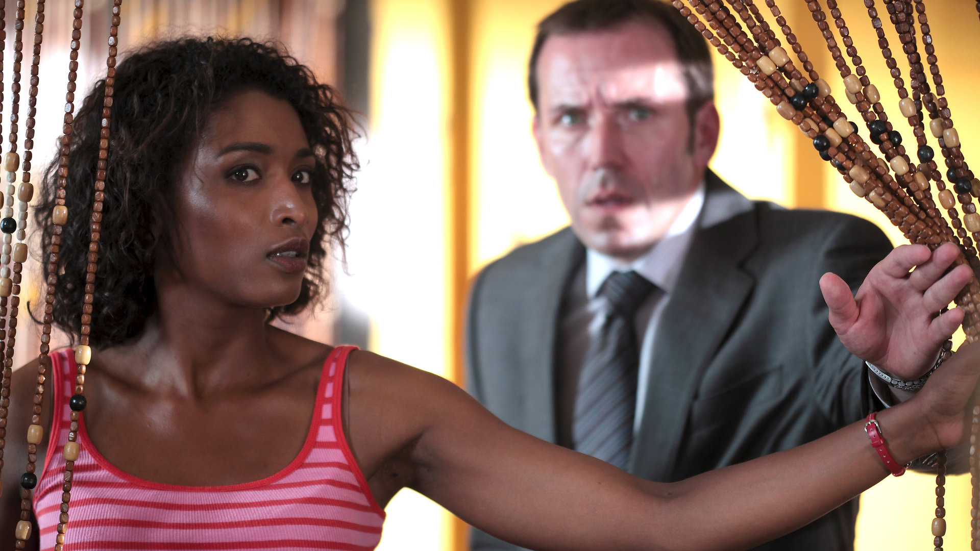 "Death in Paradise - Beste Freunde":  Camille (Sara Martins) und DI Richard Poole (Ben Miller) schieben einen Perlenvorhang zur Seite und beobachten etwas.