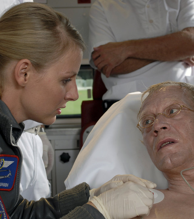 "Die Rettungsflieger - Ein großer Tag": Flieger-Oberstleutnant a.D. Karl Rottenburg (Gernot Endemann) liegt auf einer Liege in einem Krankenwagen. Sabine (Marlene Marlow) sitzt neben dem Patienten und legt ihm Elektroden an. Im Hintergrund stehen zwei Pfleger.